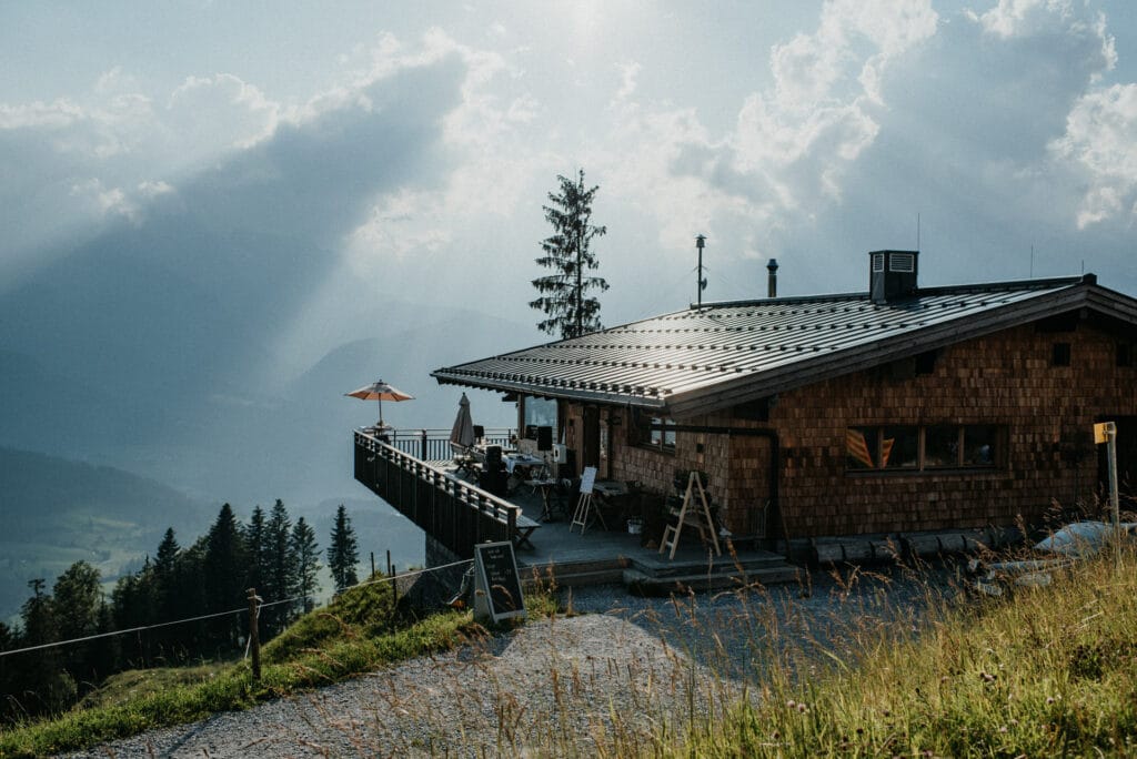 Eine rustikale Berghütte mit Holzterrasse und Blick auf ein malerisches Tal bietet die perfekte Kulisse für eine romantische Hochzeit. Sonnenlicht fällt durch die Wolken und wirft Strahlen über die Landschaft, während Stühle und Tische auf der Terrasse die Gäste zum Feiern inmitten üppiger grüner Wiesen und hoher Kiefern einladen.