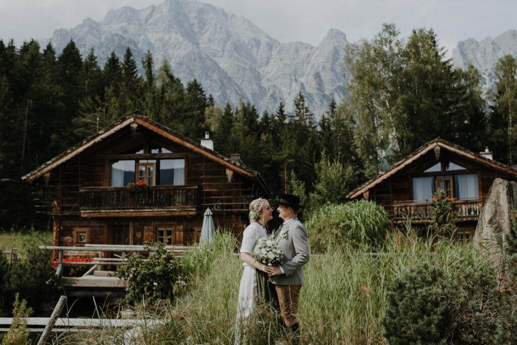 Ein Paar in traditioneller Kleidung steht umarmt da und hält einen Blumenstrauß in der Hand, was die Romantik ihrer Hochzeit verkörpert. Sie posieren anmutig vor rustikalen Holzhütten, die von hohem Gras umgeben sind. Hohe Berge und immergrüne Bäume bilden eine atemberaubende Kulisse unter dem wolkigen Himmel.