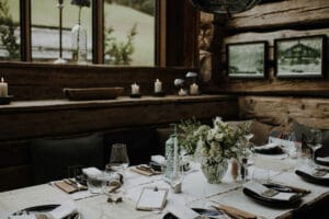 Ein rustikal gedeckter Esstisch verkörpert die Eleganz einer Hochzeit. Er besteht aus einer weißen Tischdecke, einem exquisiten Blumenschmuck, Glaswaren und gefalteten Servietten. Der Hintergrund besteht aus Holzwänden, Fenstern mit viel Grün, gerahmten Bildern und Kerzen auf Regalen – die perfekte Inspiration für die Hochzeitsplanung.