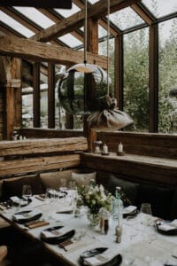 Ein rustikaler Essbereich mit Holzbalken ist perfekt für eine Hochzeitsfeier und verfügt über einen langen, für das Abendessen gedeckten Tisch. Er ist mit einem eleganten Blumenschmuck und hübsch arrangiertem Glasgeschirr geschmückt. Große Fenster sorgen für natürliches Licht und bieten einen Blick auf das üppige Grün draußen.