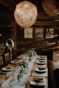 Ein rustikaler Esstisch für eine formelle Hochzeitsfeier besteht aus elegantem Geschirr und Blumenschmuck. Darüber verleiht eine große, kugelförmige, geflochtene Leuchte dem hölzernen Interieur Wärme und ist wunderschön mit gerahmter Kunst an den Wänden dekoriert. Perfekt für jeden Hochzeitsplanungstraum.