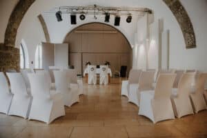 Ein kleiner, elegant eingerichteter Hochzeitsort mit Reihen weiß bezogener Stühle gegenüber einem einfachen Tisch, der mit Blumenschmuck geschmückt ist. Der Raum verfügt über gewölbte Steinwände und dezente Beleuchtung, die eine intime Atmosphäre schaffen, die perfekt für jede Hochzeit ist.