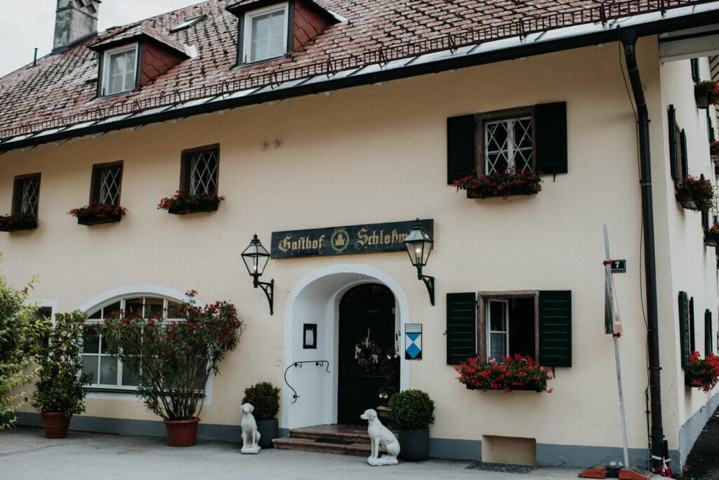 Ein malerisches gelbes Gebäude mit schrägem Dach, geschmückt mit grünen Fensterläden und Blumenkästen, bietet die perfekte Kulisse für eine bezaubernde Hochzeit. Am Eingang befinden sich zwei Laternen und zwei weiße Hundestatuen. Über der Tür steht auf einem Schild „Gasthof Schlosswirt“.