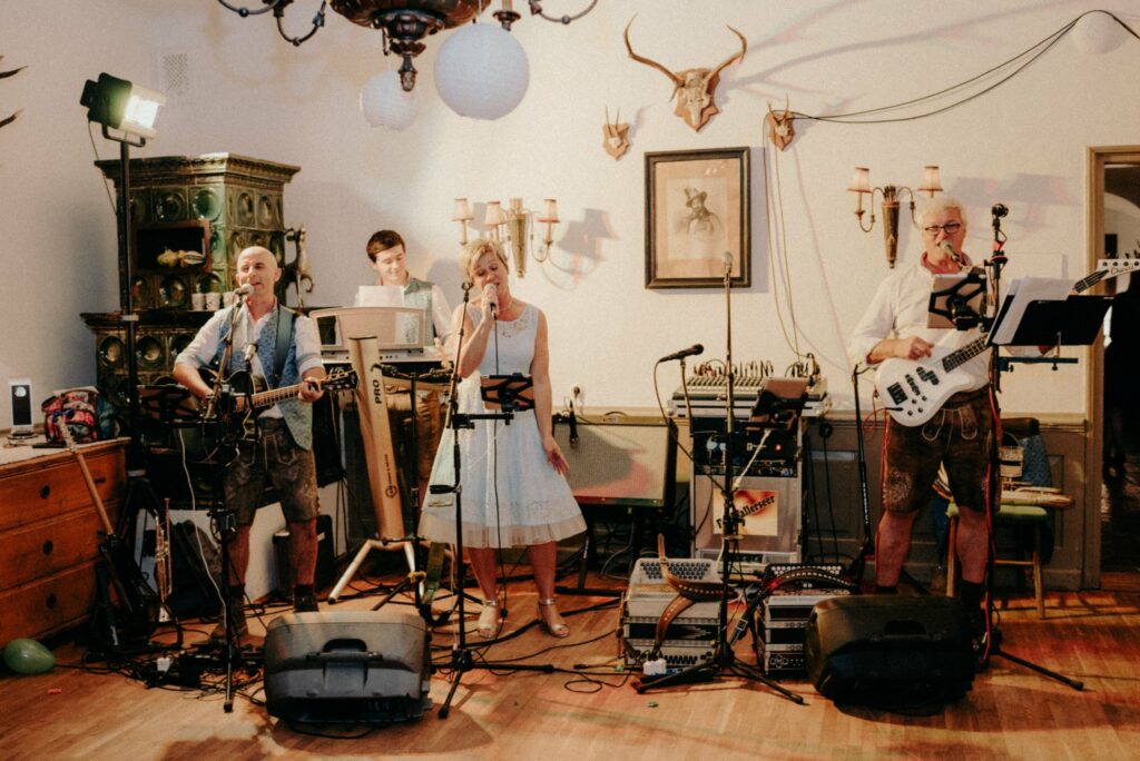 Eine Band tritt in einem gemütlichen Raum mit rustikalem Dekor auf, perfekt für eine Hochzeit. Drei Musiker in traditioneller Kleidung spielen Gitarre, Bass und singen, während ein Keyboarder im Hintergrund Tiefe verleiht. Die Kulisse besteht aus an der Wand montierten Geweihen und warmer Beleuchtung.