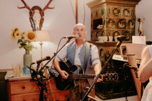 Ein Mann spielt Akustikgitarre und singt in ein Mikrofon und schafft so eine wunderbare Atmosphäre, die perfekt zu jeder Hochzeit passt. Er steht in einem gemütlichen, warm beleuchteten Raum mit einem dekorativen Ofen, Sonnenblumen in einer Vase und an der Wand befestigten Geweihen.