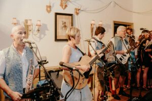 Drinnen spielt eine Band und sorgt mit einer Gitarristin und männlichen Musikern, die Trompete, Akkordeon, Bass und mehr spielen, für die perfekte Stimmung einer Hochzeit. Der mit gerahmten Kunstwerken, Hirschen und Wandleuchtern geschmückte Raum bietet eine gemütliche Atmosphäre, die ideal für Hochzeitsfeiern ist.