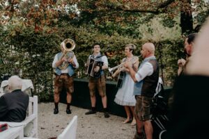 Eine Gruppe von Musikern in traditioneller Kleidung tritt im Freien auf und verleiht jeder Hochzeit eine charmante Note. Sie spielen verschiedene Instrumente, darunter Tuba, Akkordeon, Trompete und Gitarre. Die Szene spielt in einem üppigen Garten mit Zuschauern, die an Tischen sitzen. Perfekt für diejenigen, die in die Hochzeitsplanung vertieft sind.