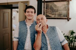 Zwei Männer lächeln drinnen. Sie tragen passende blau gemusterte Westen über weißen Hemden – perfekt für eine Hochzeit. Einer hält spielerisch einen kleinen Gegenstand vor den Mund. An der Wand hinter ihnen hängt ein Gemälde und üppige Pflanzen verleihen ihrer fröhlichen Umgebung Charme.