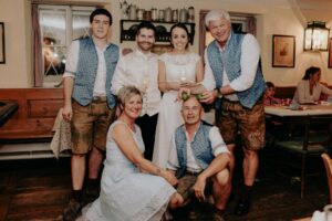 Eine Gruppe von Menschen posiert drinnen in traditioneller bayerischer Kleidung und feiert eine Hochzeit. Drei Männer, darunter der Bräutigam, tragen Westen und Lederhosen, während zwei Frauen in Kleidern, eine davon die Braut, in der Mitte stehen. Ein sitzender Mann hält eine Brezel. Die Umgebung ist gemütlich und rustikal – eine perfekte Kulisse für „heiraten“.