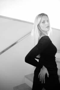 Eine Frau mit langen blonden Haaren und schwarzem Kleid posiert selbstbewusst auf einer Treppe und verkörpert Eleganz, wie geschaffen für die Hochzeitsplanung. Das minimalistische Setting und die helle Beleuchtung im Hintergrund heben ihr Profil perfekt hervor.