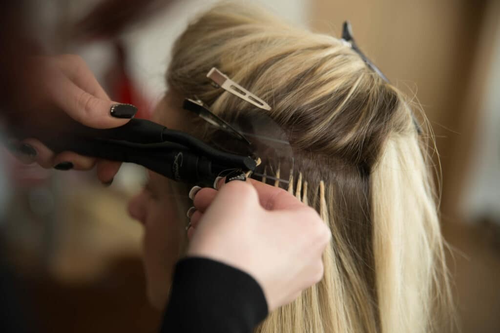 Eine Nahaufnahme zeigt einen Friseur, der einer blonden Frau sorgfältig Haarverlängerungen anbringt, möglicherweise um sie auf ihre Hochzeit vorzubereiten. Mit dunklem Nagellack und ruhigen Händen konzentriert sich der Friseur darauf, die Verlängerungen mit Klammern und einem Werkzeug an kleinen Haarpartien zu befestigen.