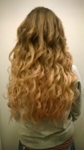 Eine Person mit langem, welligem Haar mit einem natürlichen Ombre-Effekt von Dunkelbraun bis Blond steht vor einem schlichten Hintergrund, perfekt für eine Hochzeit. Das Haar fällt in lockeren, voluminösen Locken herab und verkörpert Eleganz für jede Hochzeitsfeier.
