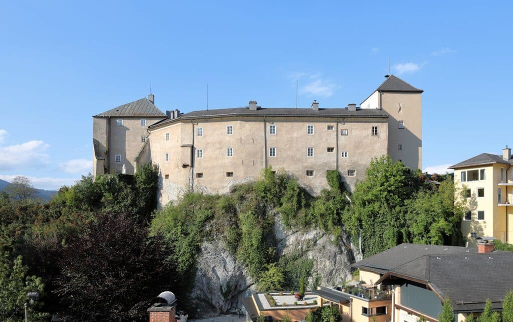 Ein großes, historisches Steinschloss thront auf einer felsigen Klippe, umgeben von Bäumen und Grünflächen, und bietet eine märchenhafte Kulisse, die sich perfekt für die Hochzeitsplanung eignet. Das Gebäude mit seinen zahlreichen Türmchen und dem schrägen Dach erhebt sich majestätisch vor dem klaren blauen Himmel. Moderne Gebäude in der Nähe sind teilweise sichtbar.