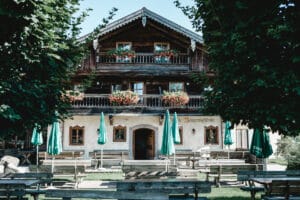 Ein charmantes, rustikales Gebäude mit Holzarchitektur und Blumentöpfen auf dem Balkon ist der perfekte Ort für eine Hochzeit. Im Vordergrund stehen Tische mit grünen Sonnenschirmen, eingerahmt von Bäumen, die an diesem sonnigen Tag eine friedliche, einladende Atmosphäre schaffen.