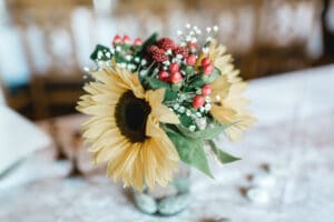 Ein bezauberndes Blumenarrangement mit Sonnenblumen, roten Beeren und grünen Blättern ziert die weiße Tischdecke und vermittelt die rustikale Eleganz, die perfekt zu einer Hochzeit passt. Der sanft verschwommene Hintergrund hebt diesen Strauß hervor und verleiht jeder Hochzeitsplanung einen Zauber.