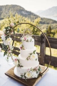 Eine dreistöckige Hochzeitstorte, perfekt für jede Hochzeit, ist mit weißen und rosa Blumen geschmückt und wird im Freien ausgestellt. Sie verfügt über einen goldenen runden Rahmen mit floralen Akzenten und einem Holzsockel. Berge und Grün dienen als atemberaubende Kulisse für dieses wesentliche Element der Hochzeitsplanung.