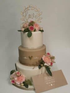 Eine dreistöckige Hochzeitstorte, perfekt für jede Hochzeit, ist mit rosa Blumen und Grün dekoriert. Die mittlere Etage ist mit einem goldenen Band umwickelt und hat ein kleines goldenes Herz. Auf der Torte steht „Liebe“ und eine Karte mit „Einladung“ ergänzt das charmante Design, ideal für alle, die schon tief in der Hochzeitsplanung stecken.