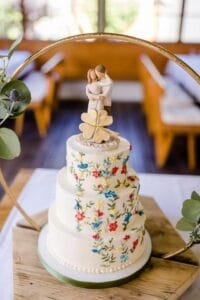 Eine dreistöckige Hochzeitstorte mit Blumenmuster und einem Brautpaar-Aufsatz ist perfekt für Ihre Hochzeit. Ein vierblättriges Kleeblatt ziert die zweite Etage. Die Torte ruht elegant auf einem Holzbrett, umgeben von Grünpflanzen vor einem goldenen Reifenhintergrund und harmoniert wunderbar mit Ihrer Hochzeitsplanung.