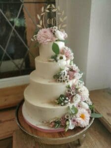 Eine vierstöckige weiße Hochzeitstorte, perfekt für eine Hochzeit, ist mit rosa und weißen Blumen, grünen Blättern und einem „Liebes“-Aufsatz geschmückt. Auf einem Holztisch mit einem Fenster im Hintergrund ist sie ein exquisites Herzstück für jede Hochzeitsplanung.