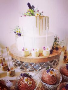 Eine zweistöckige weiße Hochzeitstorte mit goldenen Verzierungen und Blumenschmuck steht auf einem Holzständer. Um das Mittelstück herum befinden sich Cake Pops mit Blumendekorationen und Schokoladen-Cupcakes mit Sahnehaube – perfekte Details für jede Hochzeitsplanung.