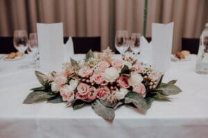 Ein wunderschön arrangiertes Blumengesteck mit rosa und weißen Rosen, umgeben von grünen Blättern, ziert eine runde weiße Tischdecke. Weingläser und gefaltete Servietten vervollständigen diese elegante Hochzeitsdekoration, ideal für alle, die gerade mitten in der Hochzeitsplanung stecken.
