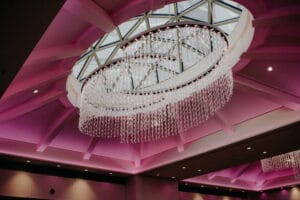 Ein großer, runder Kronleuchter mit herabhängenden Kristallsträngen hängt von einer geometrischen Decke mit einer Glaskuppel herab, perfekt für jede Hochzeit. Die Decke ist mit sanftem violettem Licht beleuchtet und schafft ein elegantes und modernes Ambiente, ideal für die Hochzeitsplanung.