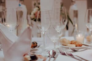 Eine Nahaufnahme eines elegant gedeckten Esstisches für eine Hochzeit, mit ordentlich angeordneten gefalteten Servietten, Weingläsern und Besteck. Brötchen und ein unscharfer Hintergrund deuten auf die sorgfältige Hochzeitsplanung für diese formelle Feier hin.