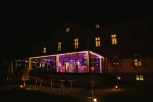 Ein großes, nachts beleuchtetes Gebäude, perfekt für eine Hochzeit, verfügt über einen modernen Bereich mit Glaswänden und farbenfroher Innenbeleuchtung. Der Außenbereich umfasst Stehtische und Stühle, dekoriert mit sanften Bodenlichtern, die eine warme Atmosphäre schaffen, ideal für eine unvergessliche Hochzeitsplanung.