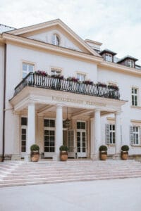 Das Bild zeigt den Eingang des Kavalierhauses Klessheim, einem eleganten Veranstaltungsort, der sich perfekt für Hochzeitsplanung eignet. Mit seiner großen Treppe, den weißen Säulen und dem mit Blumentöpfen geschmückten Balkon bietet es eine malerische Kulisse, über der stolz die Aufschrift „KAVALIERHAUS KLESSHEIM“ prangt.