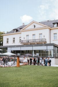 Eine Gruppe von Menschen nimmt an einer Hochzeit vor einem großen, eleganten Gebäude mit Balkon teil. Die Szene umfasst Tische mit Sonnenschirmen und grünem Rasen und lässt eine formelle Feier unter klarem Himmel vermuten – perfekt für diejenigen, die Wert auf eine sorgfältige Hochzeitsplanung legen.