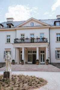 Vorderansicht eines großen, zweistöckigen Gebäudes mit heller Fassade und dunklem Dach, perfekt für Hochzeitsplanung. Es verfügt über einen markanten Eingang, Balkone und eine dekorative Statue im Vordergrund, umgeben von blühenden Büschen. Der Himmel ist klar und sonnig, ideal für Ihren Hochzeitstag.