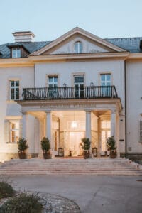 Die elegante Gebäudefassade, die sich perfekt für Hochzeitsplanung eignet, verfügt über hohe Säulen, die einen Balkon und einen beleuchteten Eingang stützen. Topfpflanzen säumen die Treppe, die zu Doppeltüren führt. Die obere Ebene verfügt über einen dreieckigen Giebel, der eine ideale Kulisse zum Heiraten unter dem sanften Abendhimmel schafft.