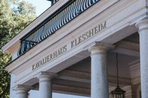 Der große Eingang des Kavalierhauses Klessheim, einem begehrten Ort für Hochzeiten, besticht durch klassische Architektur mit hohen Säulen und einem Balkon mit schwarzem Geländer. Der Name des Gebäudes ist elegant über dem Eingang eingraviert, während Bäume eine malerische Kulisse für unvergessliche Feiern bieten.