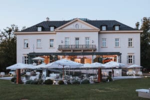Ein stattliches, zweistöckiges Gebäude mit grauem Dach und mehreren Fenstern sowie einer großen Terrasse, die sich perfekt für Hochzeitsfeiern eignet. Die Terrasse ist mit zahlreichen weißen Sonnenschirmen und eleganten Sitzgelegenheiten im Freien ausgestattet und von einem gepflegten Rasen und Bäumen umgeben.