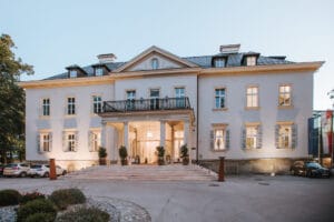 Ein großes, elegantes Gebäude mit symmetrischer Fassade, perfekt für eine Hochzeit, mit mehreren Fenstern und einem zentralen Eingang, zu dem Stufen hinaufführen. Der Veranstaltungsort ist von Bäumen umgeben und davor parken einige Autos, ideal für Ihre Hochzeitsplanung.