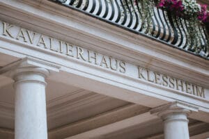 Das Bild zeigt die Fassade des Kavalierhauses Klessheim, einem perfekten Ort für Hochzeitsplanung. Es verfügt über weiße Steinsäulen und einen Balkon mit einem schwarzen Geländer und leuchtend rosa Blumen, die herabfallen.