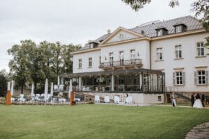 Ein herrschaftliches Anwesen mit großem Vorgarten und modernem Glasanbau eignet sich perfekt für die Hochzeitsplanung. Im Freien sind dekorierte Tische und Stühle aufgestellt, die eine elegante Hochzeit suggerieren. Laubbäume rahmen das Anwesen unter einem bedeckten Himmel ein.