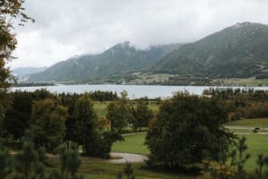 Eine ruhige Landschaft mit einem großen See, umgeben von üppigem Grün und Bergen unter einem wolkigen Himmel, bildet die perfekte Kulisse für eine Traumhochzeit. Ein gewundener Pfad führt durch die Bäume und verleiht dieser ruhigen Szene Tiefe, ideal für diejenigen, die ihre Hochzeit planen.