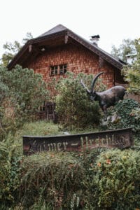 Eine rustikale Holzhütte umgeben von üppigem Grün, perfekt für intime Hochzeiten, mit einem Holzschild und einer Statue eines gehörnten Tiers im Vordergrund. Die Schindelfassade und der Kamin schaffen eine gemütliche, abgeschiedene Atmosphäre, ideal für die Hochzeitsplanung.