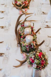 Ein langer, für eine Hochzeit geschmückter Esstisch mit Geweihen, Blumenarrangements und großen weißen Kerzen. Gläser und Besteck sind ordentlich neben jedem Gedeck angeordnet. Ein kleiner Strauß leuchtender Blumen verleiht dieser eleganten Hochzeitsfeier zusätzlichen Charme.