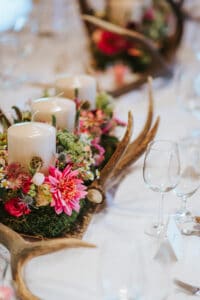 Ein atemberaubendes Tischdekor, perfekt für jede Hochzeit, mit drei großen weißen Kerzen, umgeben von rosa und roten Blumen, üppigem grünem Laub und rustikalen Geweihen. Elegant arrangierte Weingläser und Tischkarten ergänzen die Szene auf der weißen Tischdecke daneben.