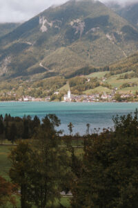 Ein ruhiger See mit türkisfarbenem Wasser erstreckt sich im Vordergrund, umgeben von üppigen grünen Bäumen – eine idyllische Kulisse, perfekt für eine Traumhochzeit. Ein malerisches Dorf mit einer markanten weißen Kirche, ideal für Hochzeiten, liegt am Fuße grüner Berge unter einem wolkigen Himmel.