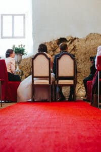 Während einer Hochzeitszeremonie sitzt ein Paar in formeller Kleidung vor einer strukturierten Steinwand. Sie sitzen elegant auf Stühlen, zu denen ein leuchtend roter Teppich führt. Eine Person in Pink sitzt an der Seite, und eine Blumenvase ist sichtbar, was die romantische Atmosphäre ihres besonderen Tages verstärkt.