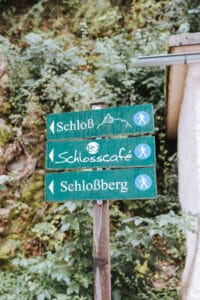 Ein grüner Wegweiser mit drei Wegweisern steht inmitten üppiger Vegetation. Das obere Schild, das nach links zum „Schloß“ mit einem Schlosssymbol führt, weist auf einen zauberhaften Ort für eine Hochzeit hin. Das mittlere zeigt zum „Schlosscafé“ und das untere führt nach rechts zum „Schlossberg“ für malerische Wandererlebnisse.