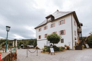 Ein altes weißes Gebäude mit roten Fensterläden steht inmitten eines Innenhofs, perfekt für Hochzeitsplanung. Auf der rechten Seite sind Treppen und im Vordergrund ein paar Tische mit Sonnenschirmen. Ein Laternenpfahl und ein Schild sind sichtbar, während grüne Bäume den Bereich unter einem bewölkten Himmel umgeben.