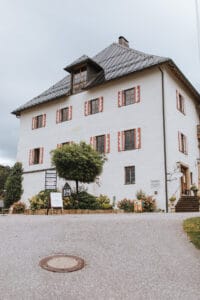 Ein malerisches, mehrstöckiges weißes Gebäude mit rot umrandeten Fenstern und einem dunklen Dach, umgeben von Grün, bietet die perfekte Kulisse für eine Hochzeit. Mehrere Schilder in der Nähe des Eingangs weisen den Gästen den Weg, während Steinstufen zur Tür hinaufführen. Der bewölkte Himmel verleiht Ihrem Hochzeitstag einen Hauch Romantik.