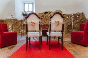 Zwei elegante Holzstühle mit Blumenschmuck auf den Rückenlehnen stehen auf einem leuchtend roten Teppich gegenüber einem kleinen Tisch. Die rustikalen Steinwände des Raums sind von rot bezogenen Sitzen und silbernen Kandelabern umgeben und eignen sich perfekt für eine Hochzeit.