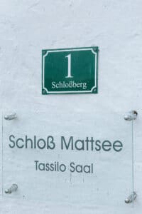 Ein Schild an der Hauswand zeigt „Schloss Mattsee“ und „Tassilo Saal“, ideal für alle, die eine Hochzeit feiern möchten. Darüber ist eine grün-weiße Tafel mit der Zahl „1“ angebracht.