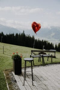 Ein roter herzförmiger Ballon ist an einem schwarzen Hocker auf einem Holzdeck befestigt und schafft eine bezaubernde Szene, die sich perfekt für jede Hochzeit eignet. In der Nähe ergänzt ein hoher Blumentopf mit roten Blumen das üppige grüne Feld, den Wald und die Berge unter einem teilweise bewölkten Himmel.