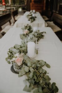 Ein elegant dekorierter Tisch verfügt über eine weiße Tischdecke, die mit einer üppigen Eukalyptusgirlande, rosa Rosen und großen weißen Stumpenkerzen in Glashaltern geschmückt ist. Perfekt für eine Hochzeit im Freien, verleihen Holzbänke dieser zauberhaften Szene Charme, die für jedes Paar geeignet ist, das davon träumt zu heiraten.